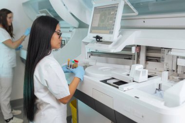 İki kadın laboratuvar teknisyeni, test tüplerini ve kan örneklerini tıbbi makinelere, otomatik dijital biyokimya analizcilerine yerleştirdi..