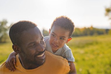Bir baba güneşli bir parkta küçük oğlunu neşeyle sırtında taşır. Doğayla çevrili bir günün tadını çıkarırlar..