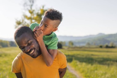 Bir baba, açık mavi gökyüzünün altındaki yeşil bir tarlada gülümseyip fısıldarken oğlunu sırtında taşır. Güneş ışıldıyor.