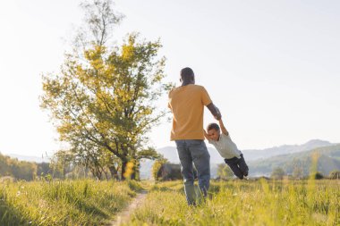 Babası neşeli küçük oğluyla dışarıda oynuyor, ellerini tutuyor ve onu daireler çizerek döndürüyor. Aile ve çocukluk mutluluk kavramı.