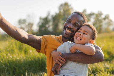 Bir baba, genç oğlunu gün ışığında çimenli bir tarlada tutarken neşeli bir şekilde gülümser ve özel bir selfie çeker..