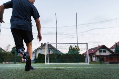 Protez bacaklı bir adam futbol kalesine doğru koşuyor. Aktif olarak alacakaranlıkta çimenli bir alanda spor yapıyor..