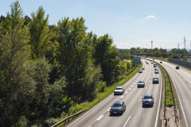 Arabalar ağaçlar ve modern altyapı ile çevrili yoğun bir otoyol boyunca ilerliyorlar. Bu sahne günlük gidip gelişleri ve şehir yaşamını gösteriyor..
