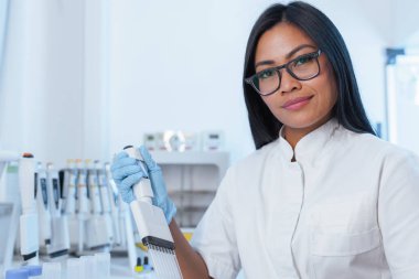 Laboratuvar önlüklü kadın bilim adamı pipetini biyoteknoloji ve tıp bilimi araştırmaları için kullanıyor. Modern bir laboratuvar ortamında çalışıyor..