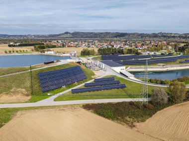 Hava görüntüsü, nehir ve tarım arazisi yakınında güneş panelleri olan bir hidroelektrik santrali gösteriyor. Bu sahne enerji üretimi etkinliğini yakalar.