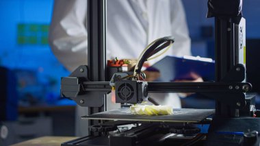 Laboratuvar önlüğü ve güvenlik gözlüğü mühendisi modern çalışma alanındaki 3D baskı sürecini denetliyor, hassasiyet ve teknolojiye odaklanıyor..