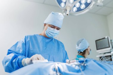 Kadın cerrah ve hemşire, temiz bir hastane odasında, parlak ışıklar altında tıbbi malzemeler kullanarak birlikte çalışıyorlar..