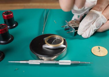 Saatçi karmaşık saat bileşenlerini eldivenli ellerle temiz bir tezgah üzerinde profesyonel ayarlarda ince aletler kullanarak birleştirir..