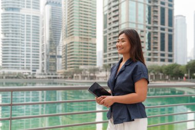 Ticari planlar üzerinde çalışmak için tablet kullanan kendine güvenen bir Asyalı iş kadınının portresi. Arka planda bir Körfez şehrinin modern gökdelenleri var..