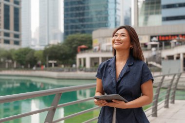 Gülümseyen Asyalı kadın girişimci elinde bir tabletle modern Körfez şehrinde güvenle yürüyor, başarı zihniyeti kavramı..