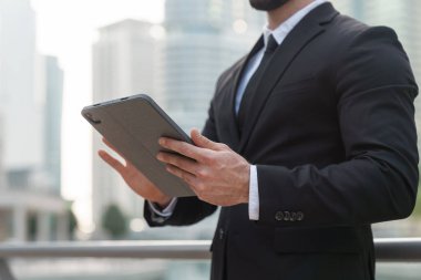 Resmi takım elbiseli bir işadamı Dubais kentsel manzarasında tableti kontrol ediyor. Gökdelenlerle çevrili..