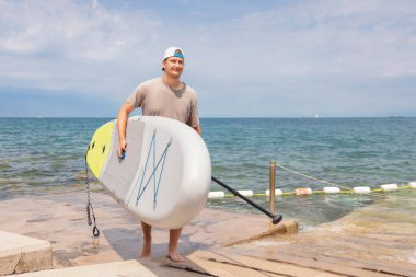 Genç adam güneşli bir yaz gününde sahilde yürürken bir raket tutuyor. Su sporlarını ve aktif bir yaşam tarzını seviyor..
