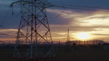 Gün batımında kırsal bir yerde trafolar ve iletim hatları olan yüksek voltajlı bir elektrik santralinin görüntüsü.