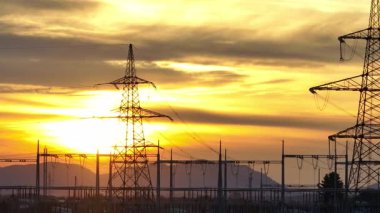 Elektrik hatları ve elektrik iletim kuleleri güneş arkalarında batarken kırsal alanda dik duruyorlar. Gökyüzü canlı renkler gösterir.