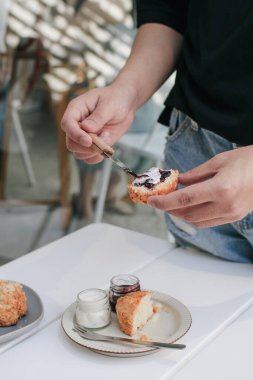 El krem peyniri ve çilek reçeli ile İngiliz çörekleri yayar, çörek genellikle buğday, arpa ya da kabartma tozu ile yulaf ezmesinden yapılan tek seferlik bir ekmektir..