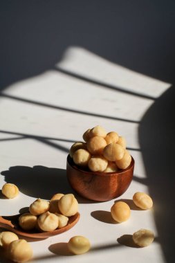 Beyaz masada, güneş ışığı ve gölgesi olan tahta kasede Macadamia fındıkları. Macadamia fındıkları sağlıklı vitaminlerle dolu bir protein kaynağıdır..