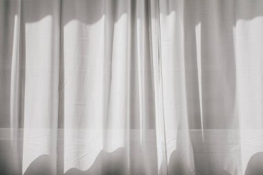 Güneş ışığı şeffaf perdenin ardından parlar. Pencerenin yanında beyaz tül sallıyordu. Sabah güneşi odayı aydınlatıyor, arka plan gölgeli..