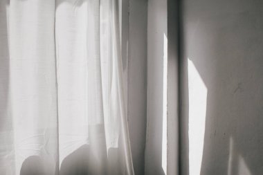 Güneş ışığı şeffaf perdenin ardından parlar. Pencerenin yanında beyaz tül sallıyordu. Sabah güneşi odayı aydınlatıyor, arka plan gölgeli..