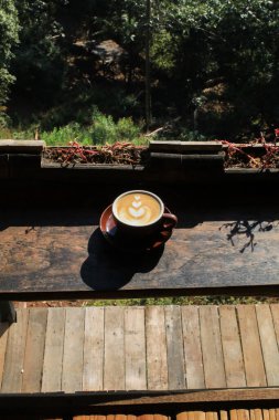 Güneş ışığı altında, ahşap masanın üzerinde latte sanatıyla sıcak kahve. Sabah kahvaltısı. Kremalı kahve fincanı aromalı..