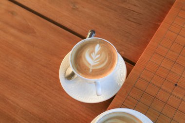 Beyaz seramik bardakta, ahşap masada köpüklü sütlü latte sanatıyla sıcak kahve. Ferahlatıcı sabah.