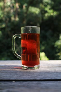 Arka planda orman olan ahşap bir masada, uzun bardakta sıcak çay. Yumuşak güneş ışığı ılık atmosferde parlıyor..