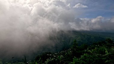 Mavi gökyüzünün altındaki yeşil dağları kaplayan bulutlar