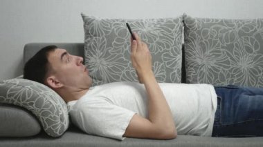 Keyifli adam gri kanepeye uzanıyor. Şaşkın adam elinde akıllı telefon tutuyor ve bir şeyden seviniyor. Kanepede akıllı telefonu olan mutlu insan.. 