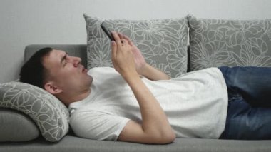 Şaşkın adam gri koltukta yatıyor. Adam akıllı telefon tutuyor ve bir şeye şaşırıyor. Kanepede akıllı telefonu olan yetişkin bir insan.. 