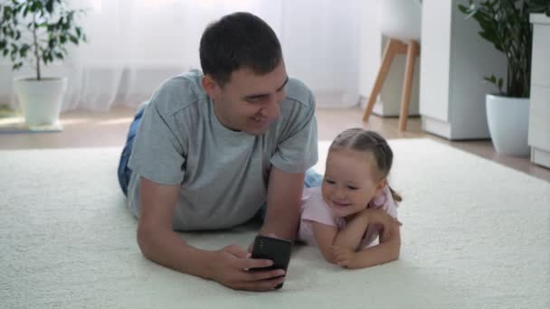 Dcera a otec se dívají na něco legračního na smartphonu a leží doma na podlaze. Malá dcera a šťastný otec spolu tráví čas. Roztomilé malé dívka mluví se svým dospělým tátou doma.