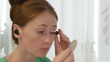 Çekici kadın kirpiklerini evde boyuyor. Kırmızı saçlı, kirpikli yetişkin bir kadın. Güzel bir insan makyaj yapar ve kulak damlasıyla müzik dinler..