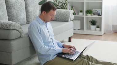 Dizüstü bilgisayar başında serbest çalışan yetişkin ya da öğrenci tipli biri. Freelancer oturma odasında yerde otururken evde çalışıyor. Beyaz adam internette bilgisayardan çalışıyor ya da okuyor.