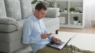 Adult man types information of credit card on laptop. Caucasian man type number of debit card on laptop. Handsome person makes online payment at home while sits on floor.