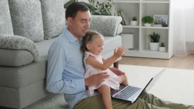 Adult father and toddler daughter listen to music on laptop and sing. Father and daughter spend time together at home. Cute small girl sits on knees of dad and and has fun with him.