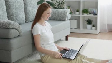 Güzel bir kadın, oturma odasında yerde otururken dizüstü bilgisayarla video görüşmesi yapıyor. Mutlu kızıl saçlı kadın evdeki bilgisayarda konuşuyor..