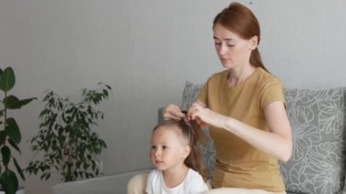Güzel anne, küçük kızı için at kuyruğu yapar. Saç stili yapma süreci. Genç anne evdeki kanepede otururken kızına saç stili yapıyor. Anne ve kızı birlikte vakit geçiriyorlar..