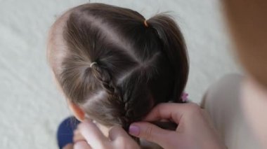 Küçük kıza örgü örme görüntüsü. Anne, küçük kızı için örgü örüyor. Kızım için saç stili yapma süreci. Örgü örme işlemi.