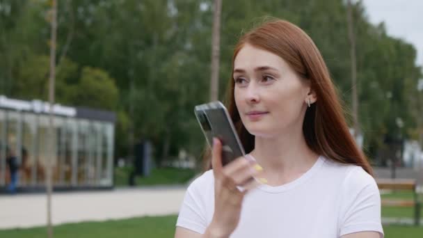 赤い髪をした美しい女性が電話に出ます 魅力的な生姜の女性は公園でベンチに座っている間 スマートフォンで話します — ストック動画