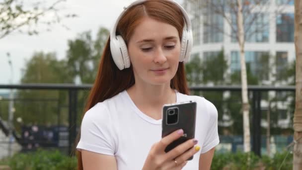 赤い髪の美しい女性は音楽に耳を傾け スマートフォンを使用しています 魅力的な女性は公園のベンチに座っている間にソーシャルメディアをサーフィンしたりニュースをチェックし ワイヤレスヘッドフォンで音楽を聴く — ストック動画