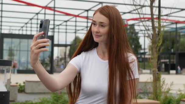 美人の生姜女がスマホで自撮り 赤い髪の魅力的な女性はスマートフォンで自撮り写真を作ります 若い女性はカメラの前でポーズし 公園のベンチに座って — ストック動画