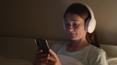 Çekici kızıl saçlı kadın akıllı telefon kullanıyor ve yatakta müzik dinliyor. Güzel, gülümseyen bir kadın gece yatak odasında akıllı telefon ve kulaklık kullanır..