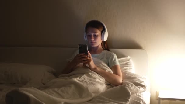 魅力的な赤い髪の女性は スマートフォンを使用してベッドの上で音楽を聴く 美しい疲れた女性は夜にベッドの上のベッドルームでスマートフォンやヘッドフォンを使用します — ストック動画