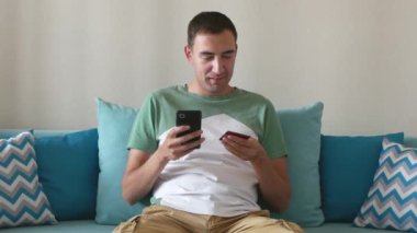 Genç adam akıllı telefondan kredi kartı bilgilerini yazar. Beyaz adam akıllı telefondaki banka kartı numaralarını yazdı. Yakışıklı adam evinde kanepede otururken online ödeme yapıyor. İnternetten alışveriş.
