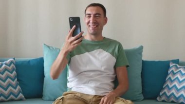 Yakışıklı genç adam video görüşmesine cevap veriyor ve akıllı telefondan konuşuyor. Gülümseyen adam evdeki kanepede oturur ve akıllı telefondan video görüşmesi yapar. Mutlu erkek arkadaşıyla konuşur..