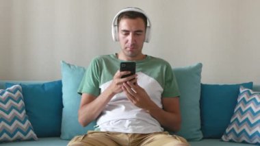 Yakışıklı genç adam akıllı telefon kullanır ve kablosuz kulaklıkla müzik dinler. Gülümseyen adam evdeki kanepede oturur ve kablosuz kulaklıkla müzik dinler..