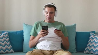 Genç adam akıllı telefondan kredi kartı bilgilerini yazar. Beyaz adam akıllı telefondaki banka kartı numaralarını yazdı. Kulaklık takan yakışıklı evde kanepede otururken internet üzerinden ödeme yapıyor..