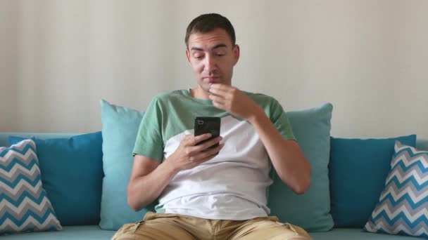 ハンサムな男は スマートフォンや笑顔で面白い何かを見ています ハッピーマンはスマートフォンを使う ハンサムな人が家でソファに座っている — ストック動画