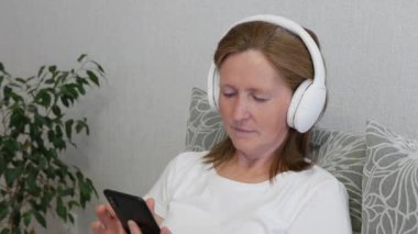 Orta yaşlı bir kadın kulaklıkla müzik dinler ve akıllı telefon kullanır. Rahat kadın evdeki kanepede oturur ve kablosuz kulaklıkla müzik dinler..