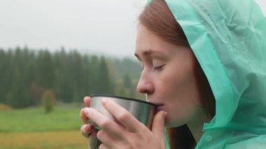 Yağmurluklu rahat kadın yağmurlu sonbahar günlerinde doğada çay içer. Kızıl saçlı çekici kadın çaydan ve köyde temiz sonbahar yağmurundan zevk alıyor..