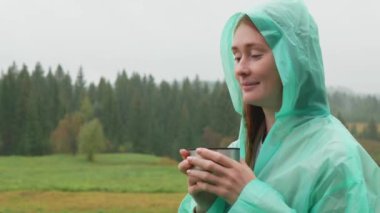 Yağmurluklu rahat kadın yağmurlu sonbahar günlerinde doğada çay içer. Kızıl saçlı çekici kadın çaydan ve köyde temiz sonbahar yağmurundan zevk alıyor..