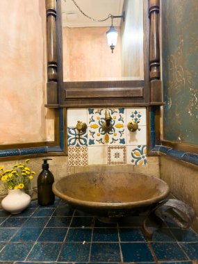 Lavabosu ve aynası olan bir banyo. Bakır ya da bronz lavabo, eski lavabo, banyoda mavi fayanslar, eski şeylerin modern banyosu. Lavabo tahtadan yapılmıştır ve üzerinde bir vazo vardır.